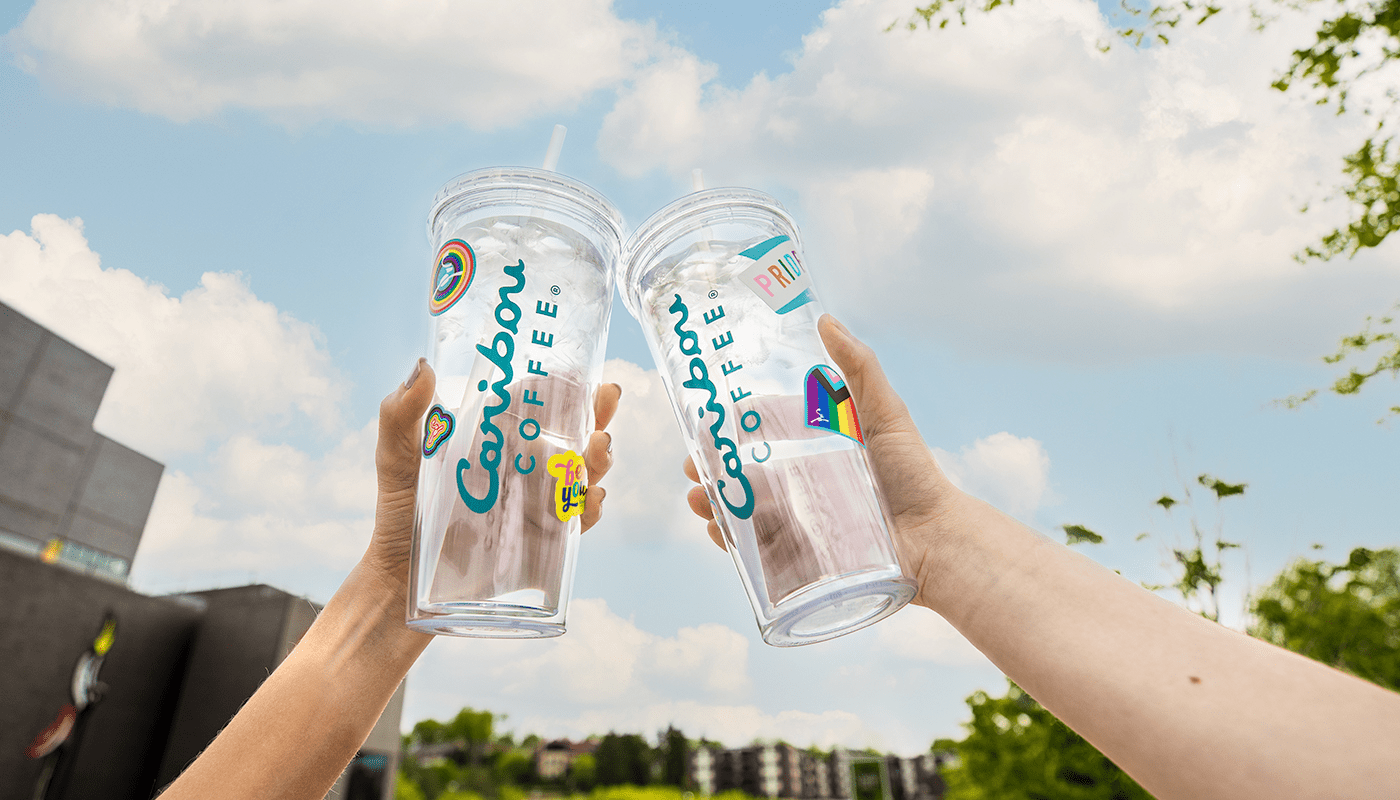 two hands holding up caribou coffee pride tumblers and giving cheers in the sky