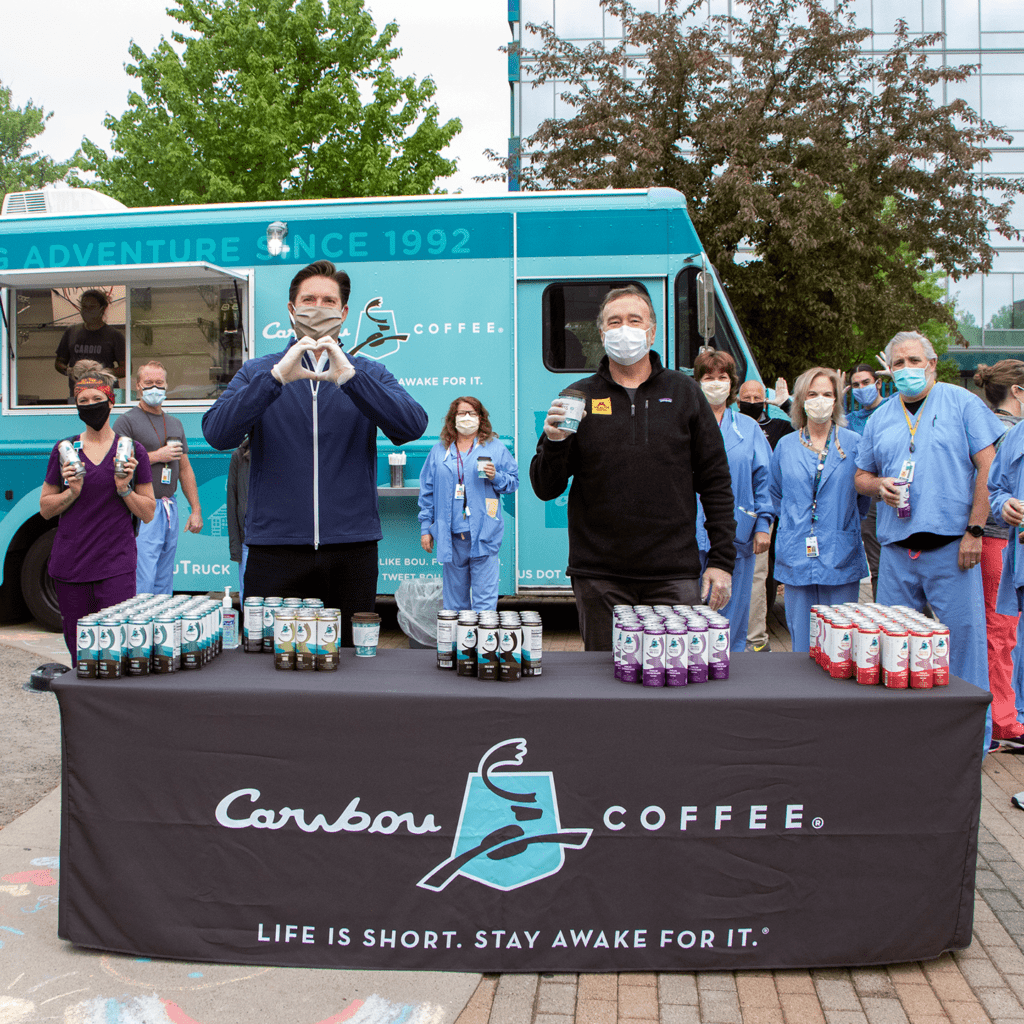 caribou coffee president and ceo john butcher making a heart-shaped sign with his hands standing behind a table of BOUsted caffeinated waters while surrounded by healthcare workers holding the beverages.