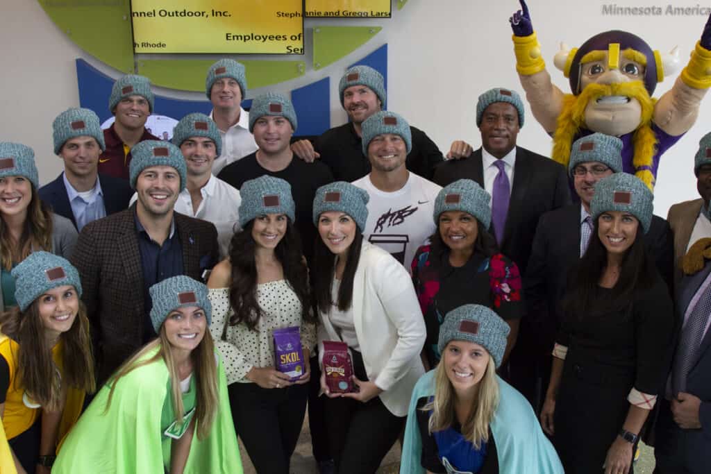 employees from the minnesota vikings, minnesota gophers, love your melon, and caribou coffee smile for a photo while wearing love your melon beanies and hold bags of caribou coffee's vikings blend and gopher blend coffees.