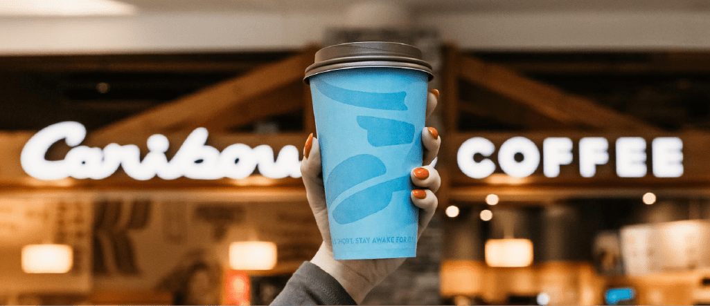 Person holding a Caribou Coffee cup against the exterior of a Caribou Coffee location