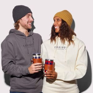 Two people wearing Caribou sweatshirts and beanies, looking at each other and cheersing with plaid tumblers