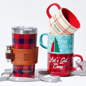 A plaid tumbler next to three festive coffee mugs stacked up