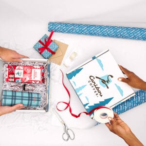 A bag of beans, a cup ornament and a teal tumbler in a box with filling, framed by wrapping supplies like scissors, tape, ribbon and wrapping paper.