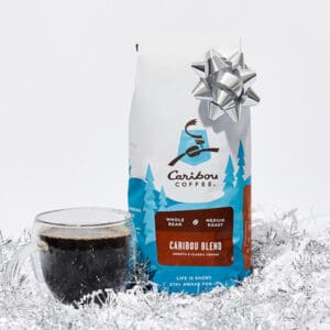 A bag of Caribou Blend coffee beans with a sliver bow on it, next to a cup of coffee in a glass mug