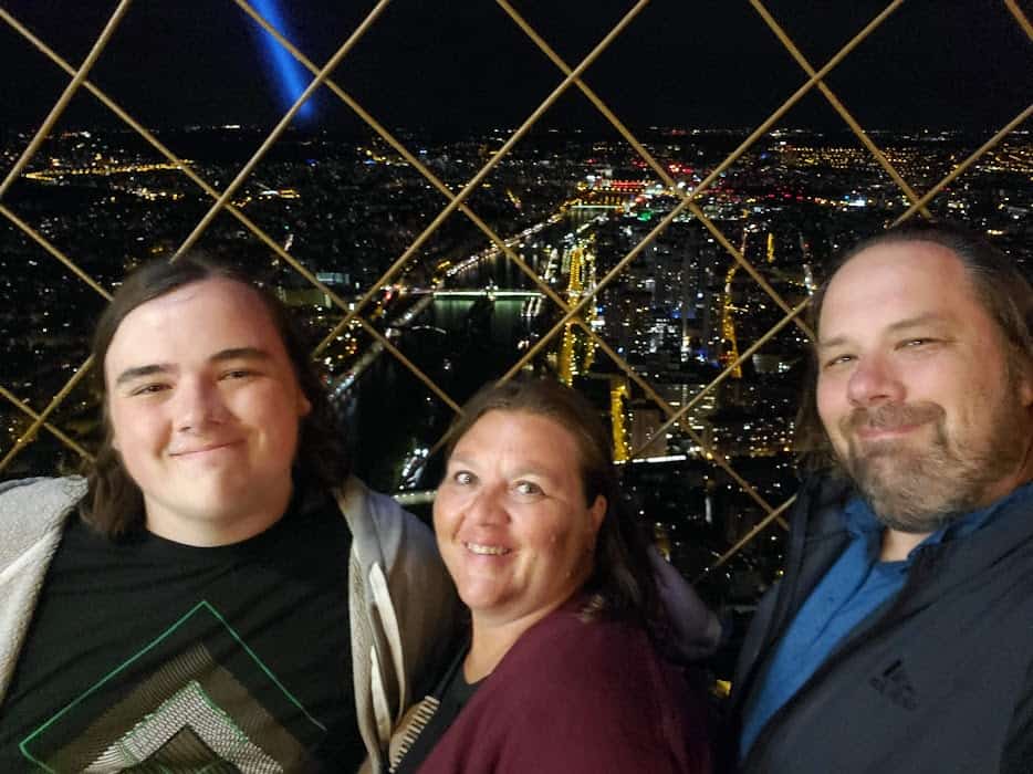 lisa's son, lisa, and her husband with a view from the top of the eiffel tower at night behind them
